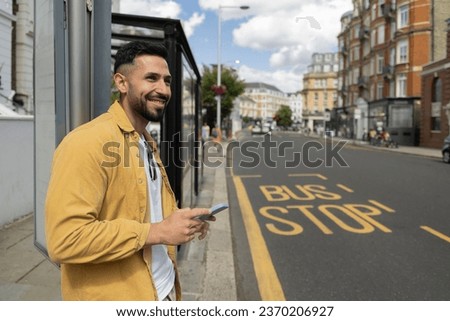 attractive young adult man waiting at the bus stop while looking around with his cell phone in his hand