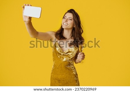 Excited woman in elegant dress taking selfie with glass of champagne posing on yellow studio background, lady making photo of herself during holiday celebration party