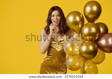 Beautiful happy european lady in golden elegant dress posing with air balloons, having birthday party over yellow background, studio shot. Excited woman having fun on holiday celebration Royalty-Free Stock Photo #2370221035