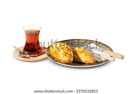 A cup of traditional Turkish tea paired with two sesame-coated pastries on a plate, accompanied by a fork and knife, set against a white backdrop