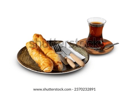 A comforting Turkish snack: two slices of borek with a glass of Turkish tea, presented on a white plate with fork and knife, all set against a white background.