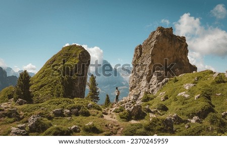 a landscape photo of an Asian female hiker trekking through a passage between 2 large rock formations spurting out of the earth with a lush green cover with mountain ranges in the distant in summer