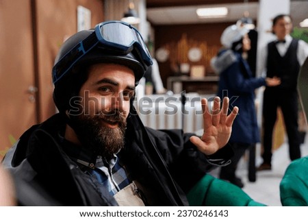 Caucasian traveler capturing winter adventure with cellphone in hotel lobby. Young man wears snow gear and takes selfie while surrounded by leisure and customers. Ski equipment suggests winter resort.