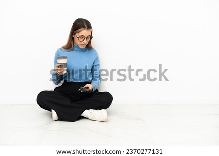 Young Caucasian woman sitting on the floor holding coffee to take away and a mobile