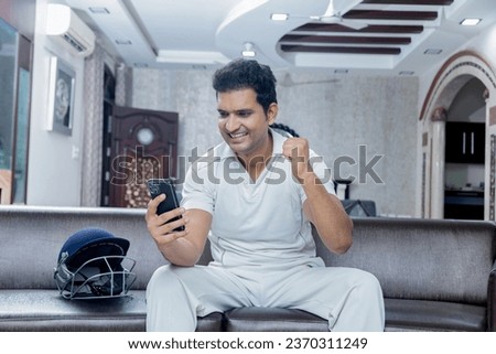 Overjoyed man in cricket dress celebrating win sitting on sofa at living room looking towards the mobile phone , Man enjoying watching cricket at home