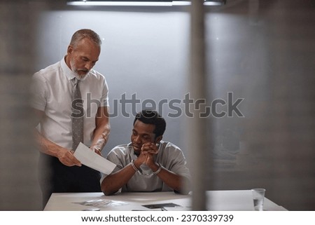 Portrait of young Black man in interrogation room with senior detective showing pictures of evidence, copy space