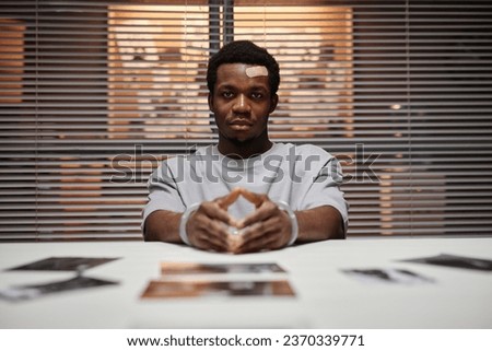 Front view portrait of Black man wearing handcuffs as criminal sitting at table in police department looking at camera with smug face expression, copy space