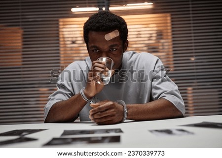Portrait of Black man wearing handcuffs as criminal sitting at table in police department and drinking water, copy space