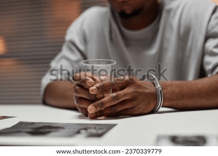 Close up of anonymous black man wearing handcuffs sitting at table in police department and holding glass of water, copy space