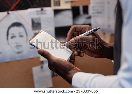 Closeup of detective writing in notebook while investigating case in office standing by evidence board, copy space