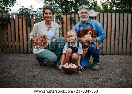 Grandmother, grandfather and toddler holding chicken and collected eggs while smiling for the camera in the garden