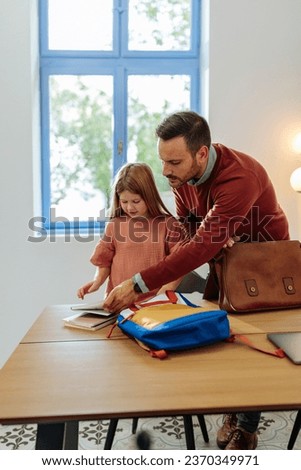 A Caucasian father and daughter are standing in a modern dining room, the father reaching over her school books