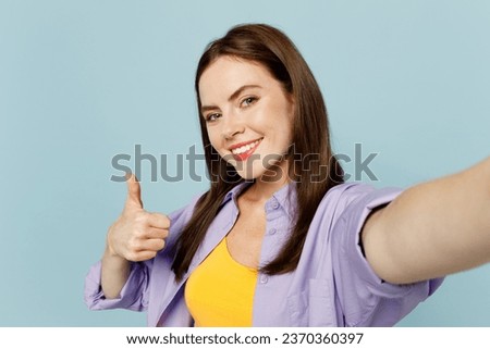 Close up young happy woman wears purple shirt yellow t-shirt casual clothes doing selfie shot pov on mobile cell phone show thumb up isolated on plain pastel light blue background. Lifestyle concept