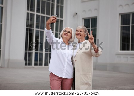 Beautiful Senior women taking selfie photo on mobile phone at city, smile against background of white building. Two older female friends on vacation.