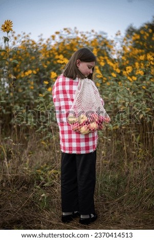 8-Year-Old Girl in Red and White Checkered Coat Among Yellow Flowers with Eco Bag with apples