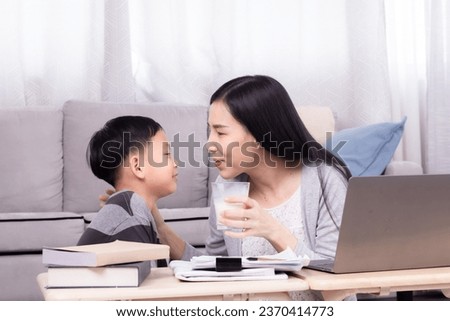 Side view Asian boy kid give mom glass of milk with love tenderness care, son bring cup of milk to mother and sit together while female working on technology laptop in living room happy mom lifestyle Royalty-Free Stock Photo #2370414773