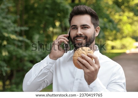 Lunch time. Young businessman with hamburger talking on smartphone in park