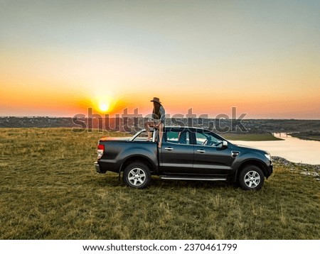 girl sitting on the roof of the pick up truck on the edge of cliff above a river and a samll village on the backgorund during sunset Royalty-Free Stock Photo #2370461799
