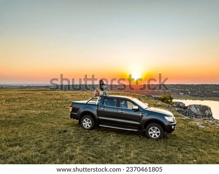 girl sitting on the roof of the pick up truck on the edge of cliff above a river and a samll village on the backgorund during sunset Royalty-Free Stock Photo #2370461805