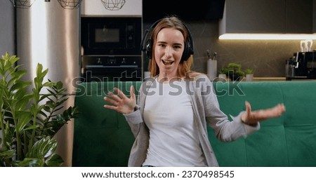 Cheerful young woman wearing wireless headphones, listening music, dancing sitting on sofa, enjoying free movement. Girl listen song in head phone at home kitchen background