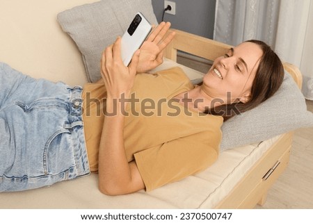 Device addiction. Web browsing. Cellphone usage. Resting with technology. Relaxed smiling brown haired woman wearing beige T-shirt lying on sofa holding smartphone in living room.