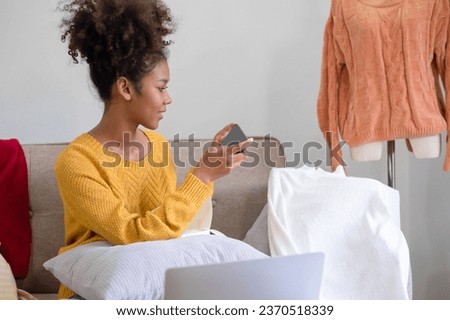 Young female business owner in the standing near clothesline with cardboard boxes in workplace taking selfie with clothes, product using smartphone to post and sell in online website. 
