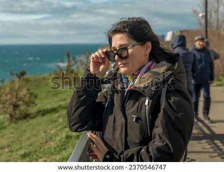 A middle-aged woman in dark glasses, dressed in a winter jacket, stands on the embankment and looks towards the sea. Time of year - off-season, spring, autumn.
