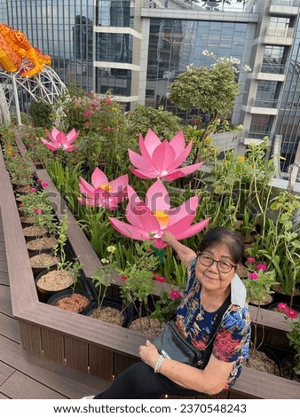old grandma posing with lotus flowers. 82 year old grandma is always healthy, and always cheerful. wearing flowery clothes and wearing glasses. stylish like a young person. photo on top of a building 