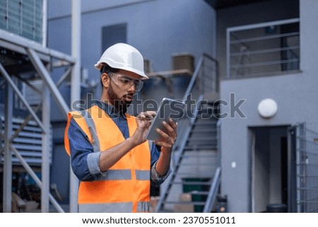 Serious thinking and focused engineer working in factory in hard hat and vest, man using tablet computer reading diagram, and checking equipment, inside industrial factory. Royalty-Free Stock Photo #2370551011