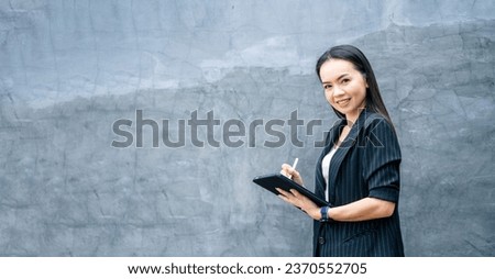 Portrait Asian businesswoman professional in black business suit using a digital tablet standing in the office building representing business oriented concept. Business stock photo.
