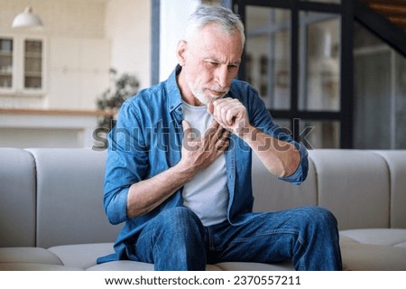 Mature man suffering from dry cough and chest pain while sitting alone on sofa in living room. Male feeling sick, has fever and respiratory infection symptoms. Healthcare concept Royalty-Free Stock Photo #2370557211