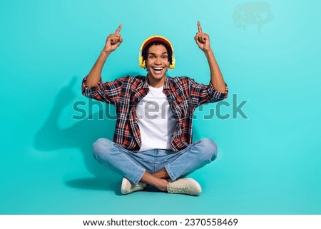 Full size portrait of handsome man sit floor listen music headphones direct fingers up empty space isolated on turquoise color background