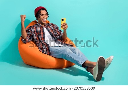 Full length photo of overjoyed ecstatic guy wear hat sit on bean bag hold smartphone win gambling isolated on turquoise color background