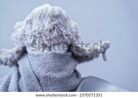 A frozen man all bundled up in a fur trappers hat, scarf, and parka, covered in snow and frost trying to stay warm on a very cold gray Winter's day. Royalty-Free Stock Photo #2370571351