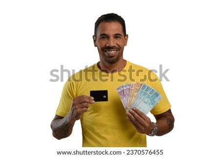 man holds a black credit card and Brazilian money, wearing a yellow shirt on a white background