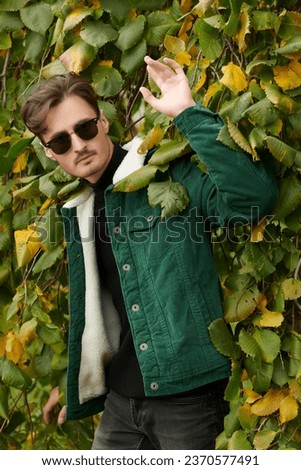 Portrait of a handsome brunette man in a green corduroy jacket and black sunglasses standing among autumn foliage in the park. Autumn men's fashion.