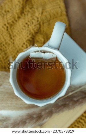 Mug of tea on a book with a mustard yellow knitted blanket. Boho grass and tea bag. 