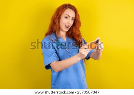 Nice addicted cheerful young red-haired doctor woman over yellow studio background using gadget playing network game