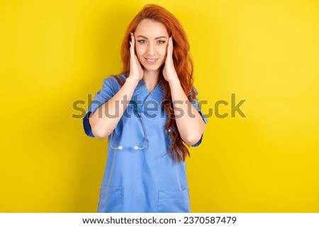 Happy young red-haired doctor woman over yellow studio background touches both cheeks gently, has tender smile, shows white teeth, gazes positively straightly at camera,