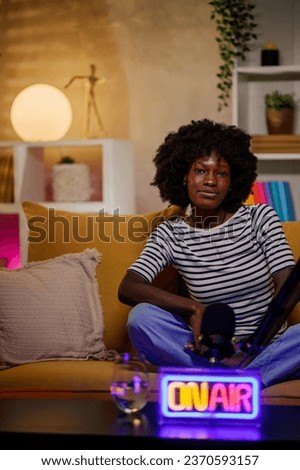 Vertical shot of a portrait of an african american radio host recording podcast in a home broadcasting studio while sitting on a yellow sofa and looking into the camera. Live stream concept.