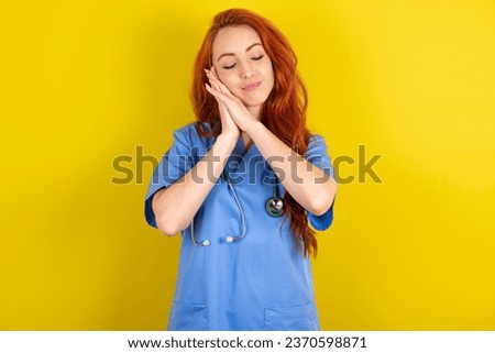 young red-haired doctor woman over yellow studio background leans on pressed palms closes eyes and has pleasant smile dreams about something