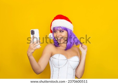 A young happy smiling caucasian woman with purple hair in white festive corset, red Santa Claus hat  takeing a selfie with her mobile phone isolated on color yellow background. Christmas and New Year