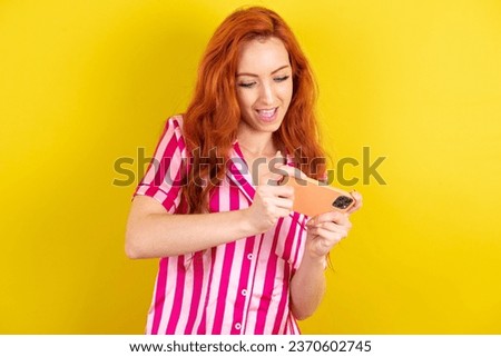 Young red haired woman wearing pink pyjama over yellow studio background holding in hands cell playing video games or chatting
