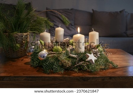 First Advent, wreath with one burning white candle and Christmas decoration on a wooden table in front of the couch, festive home decor, copy space, selected focus, narrow depth of field Royalty-Free Stock Photo #2370621847