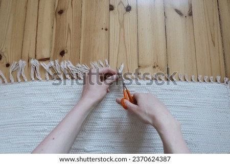Close-up image of woman cutting the fringes of a beige rug with orange scissors. Wooden floor, natural white light from a window. Half of the fringes long, half of fringes are already cut. Fresh home Royalty-Free Stock Photo #2370624387