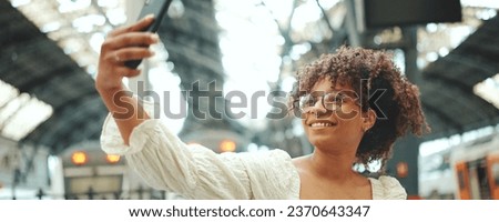 Young woman in glasses sits at the station and takes a selfie. Positive woman using mobile phone outdoors in urban background.