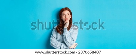 Thoughtful cute woman with red hair, looking upper left corner logo and thinking, imaging something, standing over blue background.