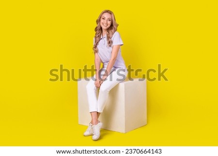 Full size photo of gorgeous positive girl wear violet polo white trousers sit on cube platform isolated on yellow color background