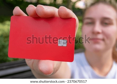A girl on the street is holding a red credit card with no writing on it. A close-up of a red bank card in her hand. Bank card chip.  Royalty-Free Stock Photo #2370670517
