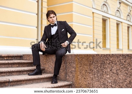 Serious handsome guy in tuxedo sitting on stairs, strong looking at camera. Confident businessman young man in tux posing at urban street outdoors, luxury image James Bond style. Copy ad text space Royalty-Free Stock Photo #2370673651